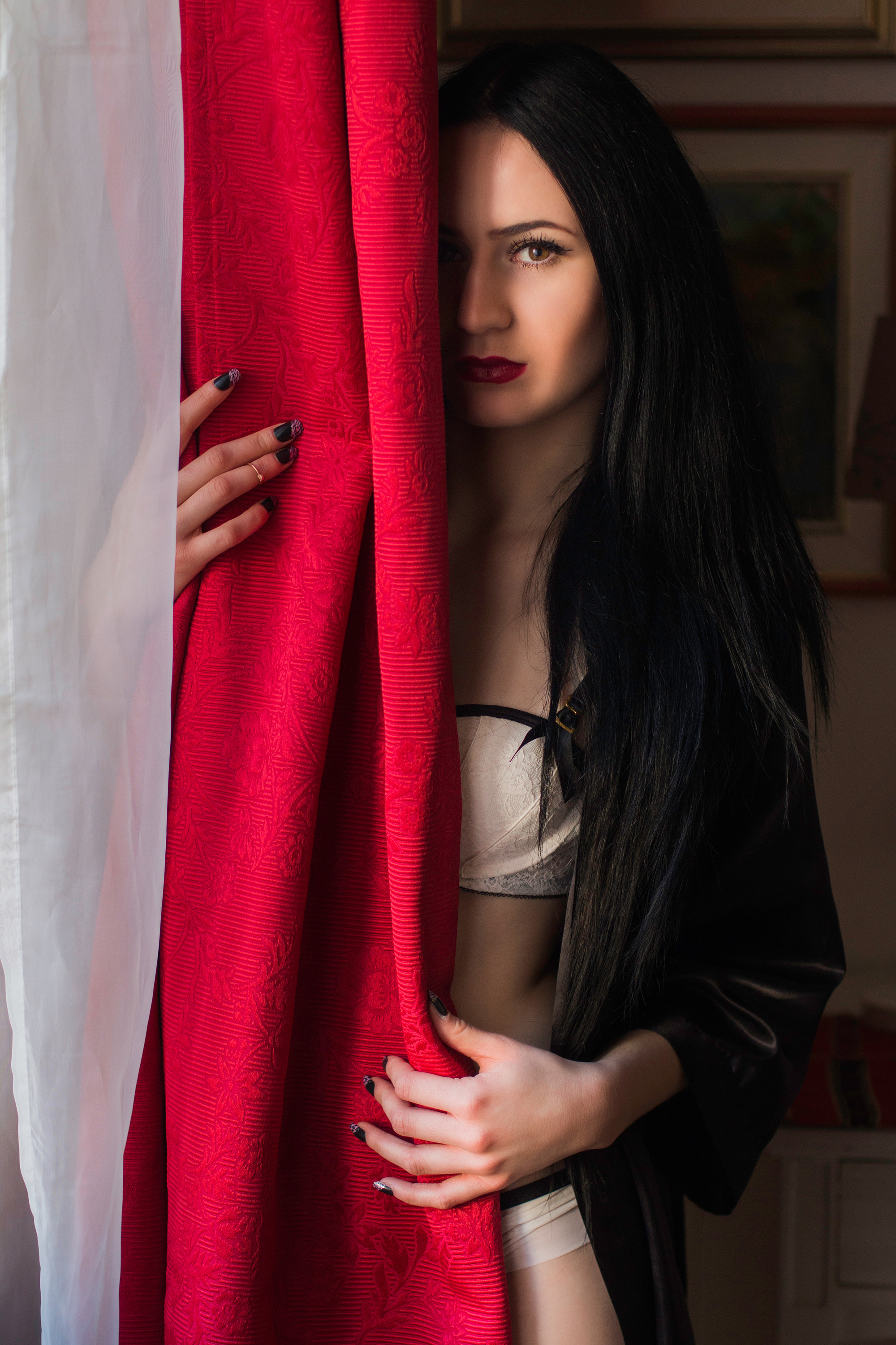 Beautiful young girl in kimono hiding behind the red curtains in the interior near window. Sexy shy lady.